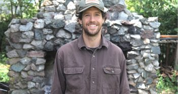 A man in a hat standing in front of a stone fireplace.