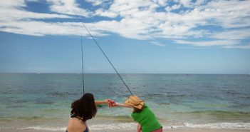 Two women sitting on a beach with a fishing pole.