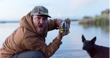 A man in a hat holding a fish and a dog.