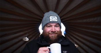 A bearded man holding a mug in a yurt.