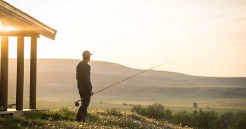 A man standing in front of a cabin with a fishing rod.