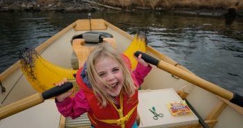 A young girl in a yellow life jacket paddles a canoe.