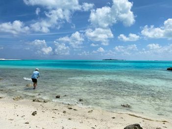 A man walks along a beach with clear blue water.