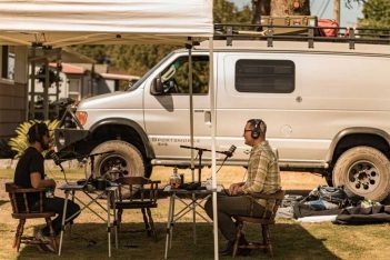 Two men sitting under a tent in front of a van.