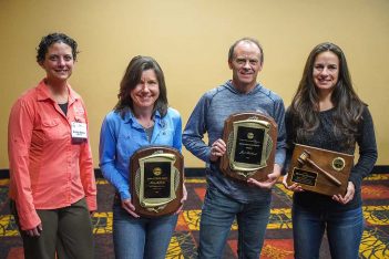 Four people holding plaques in front of a room.