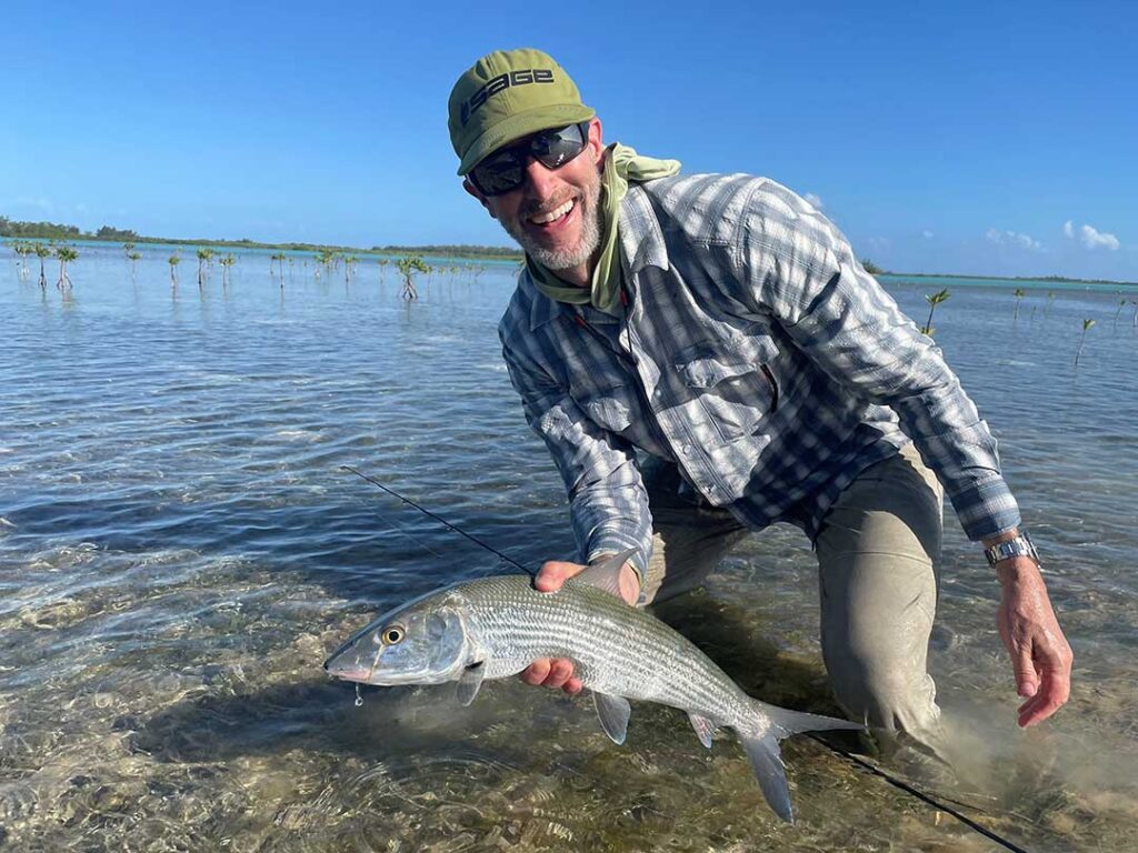 A person wearing sunglasses and a hat kneels in shallow water, holding a large fish with both hands. The clear water and a blue sky are visible in the background.