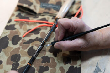 A person is carefully applying paint to a fishing rod resting on a camouflage-patterned fabric.