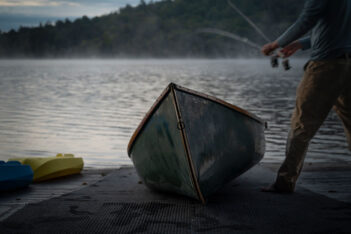 A person holding fishing rods stands on a dock next to a small boat with mist rising from a lake and forested hills in the background.