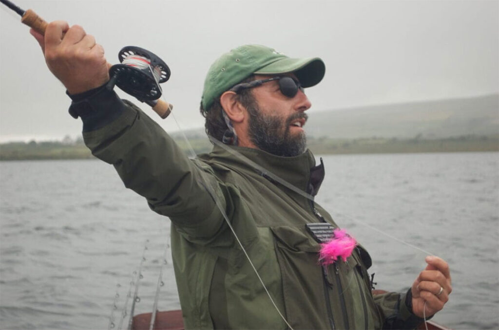 A bearded man in a green jacket and cap holds a fishing rod while standing on a boat in a foggy lake.