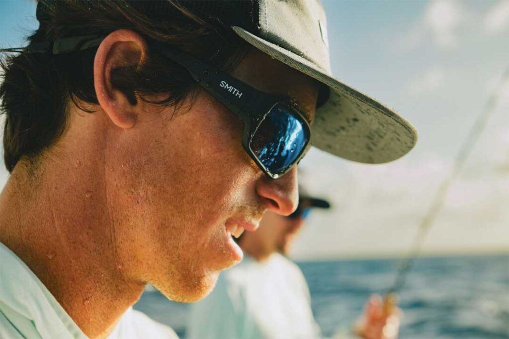 A person wearing a hat and sunglasses is looking intently to the side, with another individual blurred in the background, onboard a boat with the ocean visible.