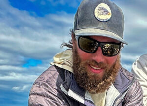 A man wearing a cap and sunglasses smiles at the camera with a blue sky and clouds in the background.