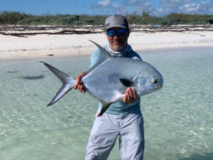 Person in light blue attire and sunglasses holding a large fish while standing in shallow clear water on a beach.