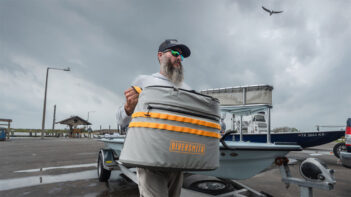 A person with a beard and sunglasses carries a gray, yellow-striped Riversmith bag near a boat trailer on a cloudy day. A bird flies in the sky above.