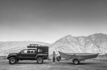 A woman stands between a black truck with a camper and a covered boat on a trailer parked in a scenic mountainous area.