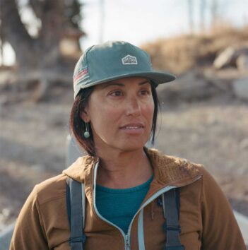 A woman wearing a green cap, brown jacket, and teal shirt looks off camera in an outdoor setting.