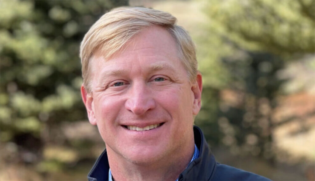 A man with short blond hair smiling outdoors with trees in the background.