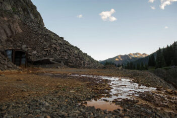 A rocky mountainside with debris, an old wooden structure, and a shallow stream of water with a distant view of snow-capped mountains and pine trees under a clear sky.