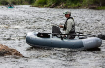 A person fishing from an inflatable boat on a flowing river, wearing a hat and sunglasses.