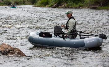 A person fishing from an inflatable boat on a flowing river, wearing a hat and sunglasses.