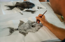 A person uses a fine brush to paint a detailed image of a fish on a white sheet, with another painted fish image in the background on a napkin.