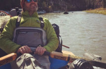 A person in fishing gear, wearing sunglasses and a beanie, relaxes on a boat floating down a scenic river with forested hills in the background.