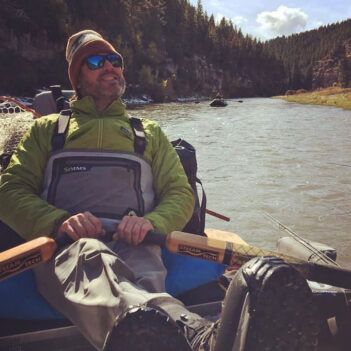 A person in fishing gear, wearing sunglasses and a beanie, relaxes on a boat floating down a scenic river with forested hills in the background.