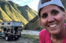 A woman in a pink shirt and white cap smiles in front of a truck with a pop-up camper in a mountainous area.