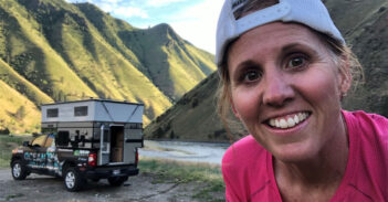 A woman in a pink shirt and white cap smiles in front of a truck with a pop-up camper in a mountainous area.