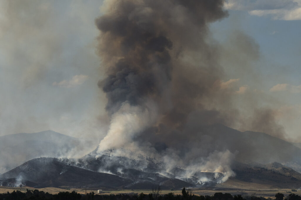 Thick smoke billows from a wildfire consuming a hilly landscape, with flames visible at the base and a dense plume rising into the sky.