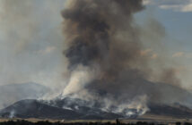 Thick smoke billows from a wildfire consuming a hilly landscape, with flames visible at the base and a dense plume rising into the sky.