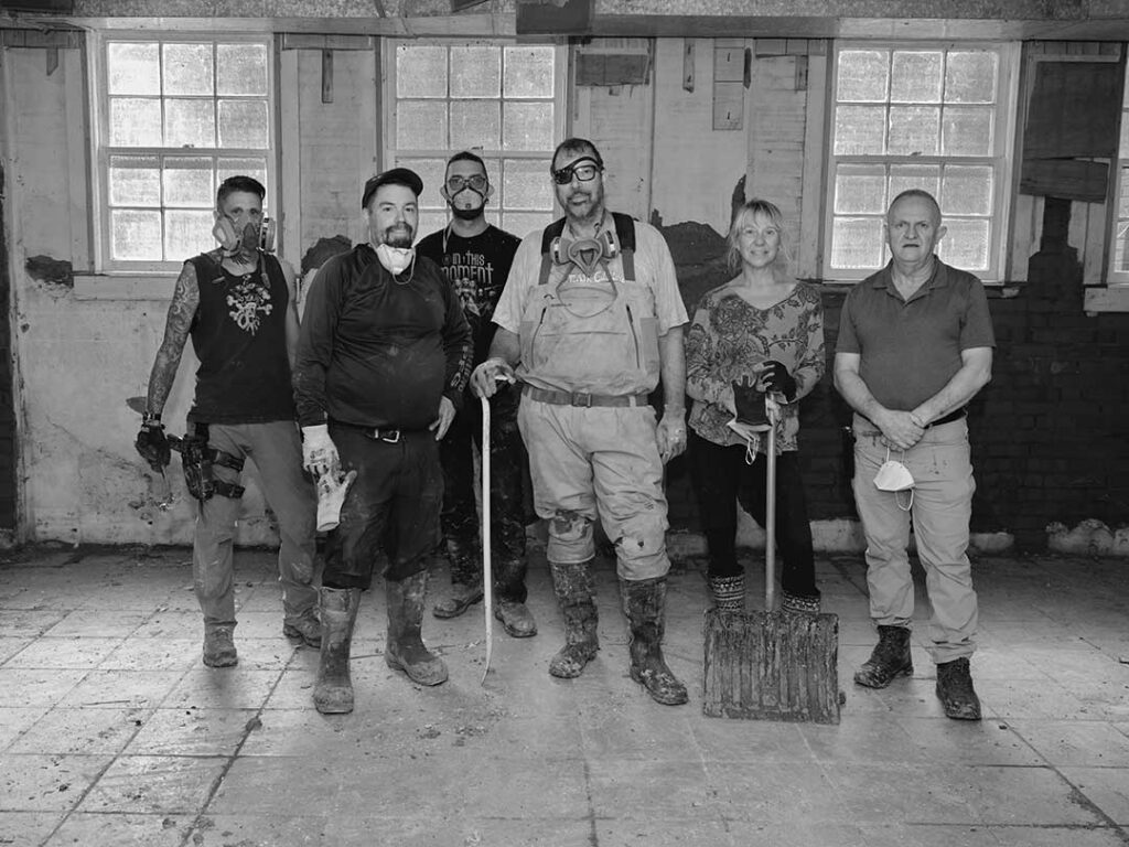 Six people stand in a row inside a building under renovation, wearing work clothes and holding tools, amid a partially tiled floor and windows in the background.