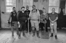 Six people stand in a row inside a building under renovation, wearing work clothes and holding tools, amid a partially tiled floor and windows in the background.