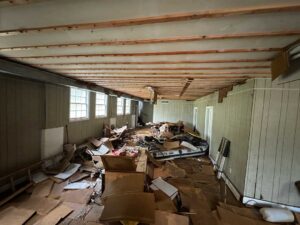 An abandoned room with scattered debris and boxes on the floor. The ceiling has exposed wooden beams, and the walls are painted light gray.