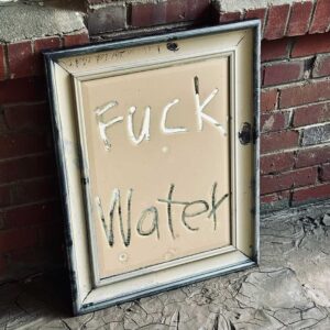 A framed board leans against a brick wall, displaying the handwritten phrase "Fuck Water.