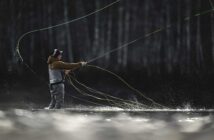 Person fly fishing in a river, wearing a jacket, cap, and waders, surrounded by splashes of water.