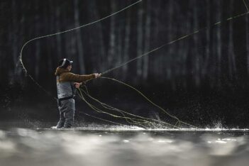 Person fly fishing in a river, wearing a jacket, cap, and waders, surrounded by splashes of water.