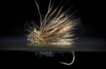 Close-up of a fishing fly with a barbed hook, featuring realistic feathery textures and intricate details, set against a dark background.