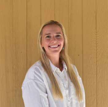 A person with long blonde hair smiles and stands in front of a wooden wall.