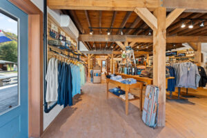 Clothing store interior with wooden beams, racks of shirts, tables displaying clothes, and a suitcase. Large windows let in natural light.