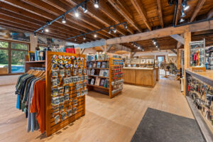 A store interior with wooden beams, clothing racks, books, and various merchandise on display. Bright lighting illuminates the space.