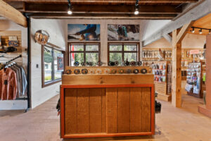 A wooden display showcasing fishing reels inside a rustic store. Clothing is on hangers to the left, and fly fishing gear is on shelves to the right. Large windows provide natural light.