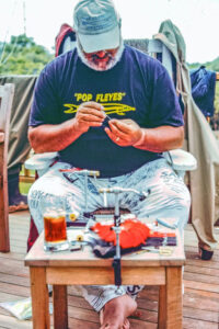 A bearded man sits on a deck tying a fishing fly with tools and materials on a small table. He wears a "Pop Fleyes" shirt and cap, with clothes hanging in the background.