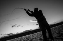 Silhouetted person casting a fishing line in a river during sunset.