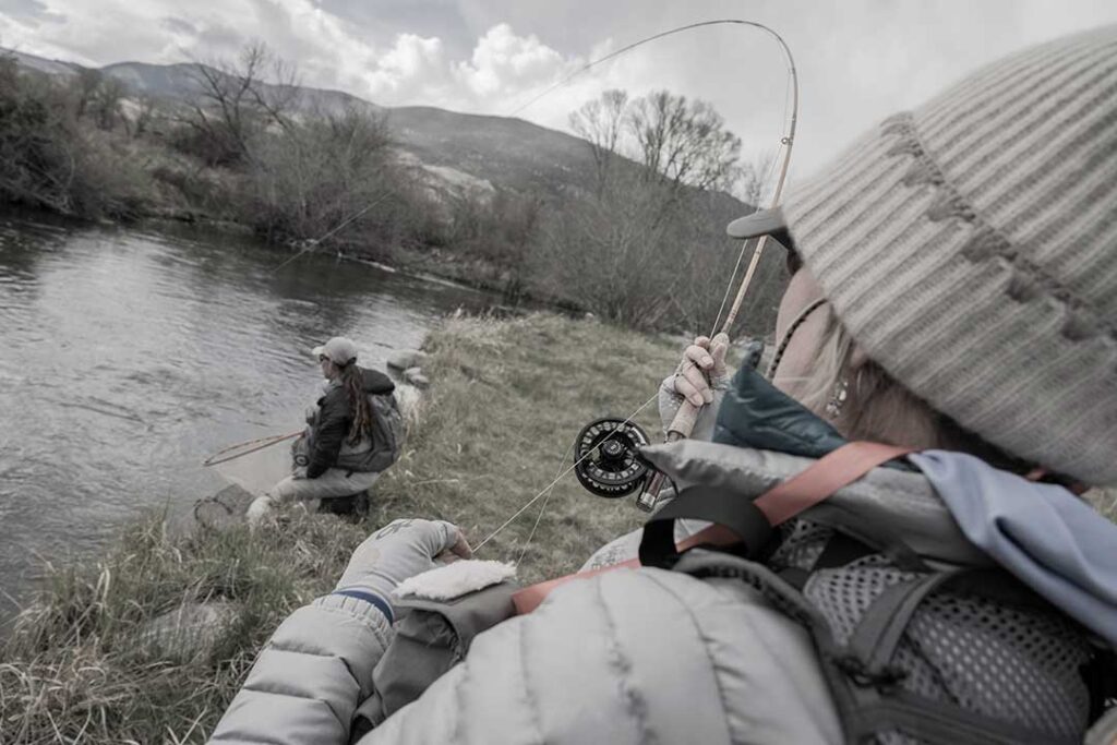 Two people are fly fishing by a river. One person holds a fishing rod in the foreground, while the other walks near the riverbank. Overcast sky and mountains are visible in the background.