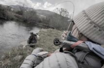 Two people are fly fishing by a river. One person holds a fishing rod in the foreground, while the other walks near the riverbank. Overcast sky and mountains are visible in the background.