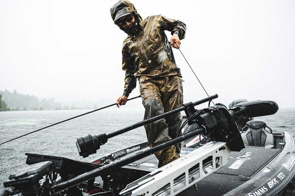 Person in rain gear stands on a boat holding a rope, surrounded by water under a cloudy sky.