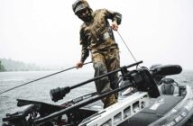 Person in rain gear stands on a boat holding a rope, surrounded by water under a cloudy sky.