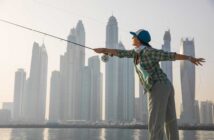 A person in a plaid shirt and cap casts a fishing line, standing near water with a city skyline in the background.