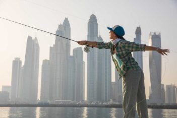 A person in a plaid shirt and cap casts a fishing line, standing near water with a city skyline in the background.