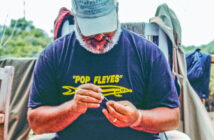 Man sitting outdoors, wearing a cap and a "Pop Fleyes" t-shirt, focused on tying a fishing fly.